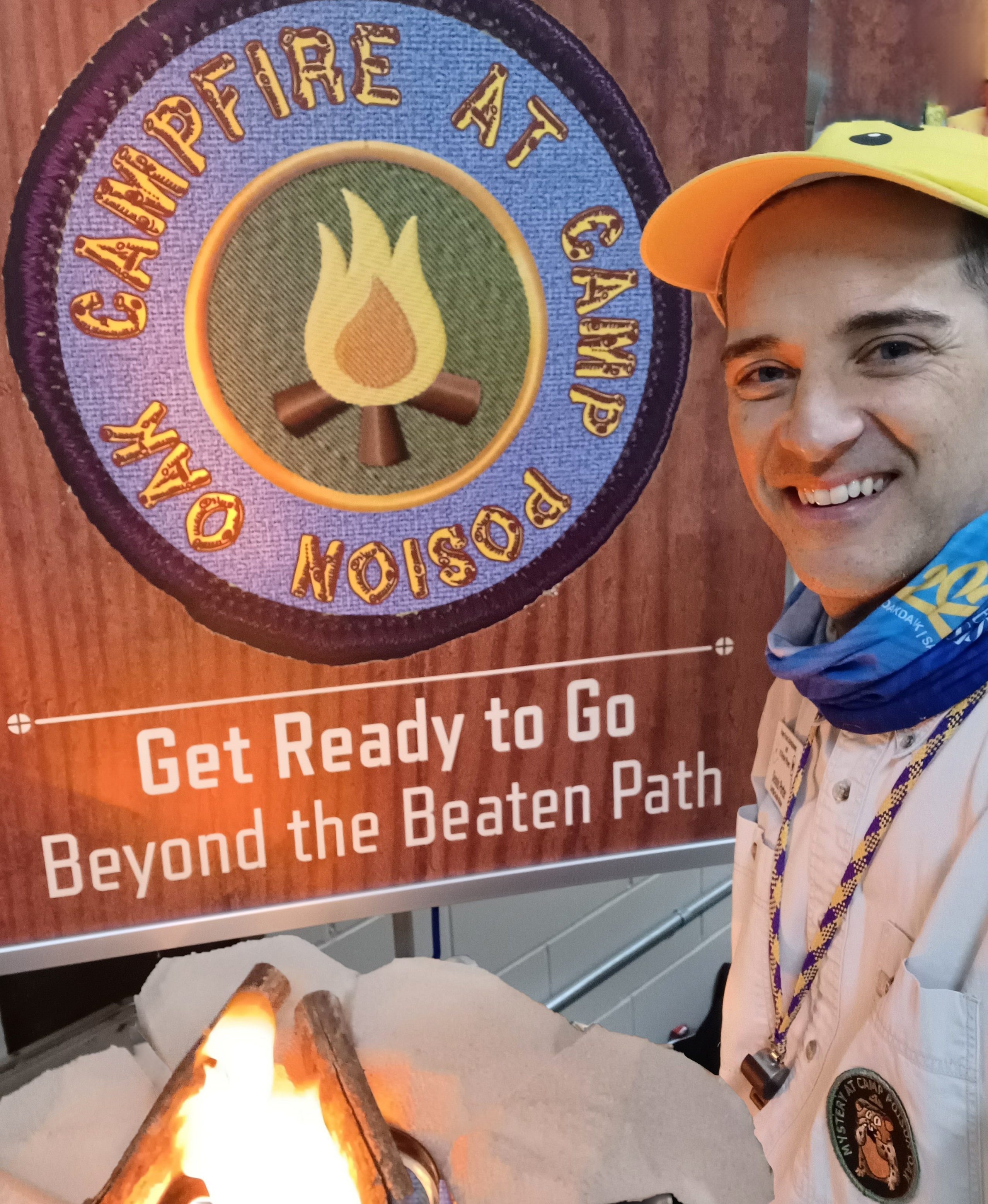 Photo of Dennis Porter dressed in camping clothes with campfire in front of sign that reads "Campfire at Camp Poison Oak. Get Ready to Go Beyond the Beaten Path."
