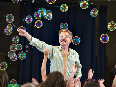 Man waving a wand with bubbles flying all around him in front of young kids