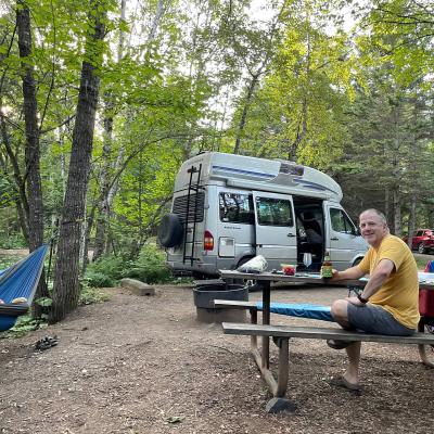 A man sits at a picnic table in front of a van in wooded area. Another person is in a nearby hammock.