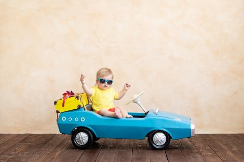 Toddler wearing sunglasses sitting in a toy car with a suitcase on the back