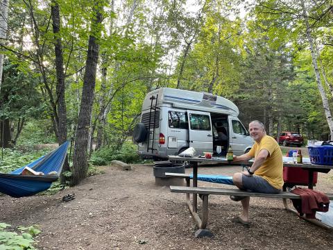 A man sits at a picnic table in front of a van in wooded area. Another person is in a nearby hammock.