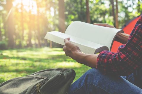 Person reading a book near a forest with a backpack nearby