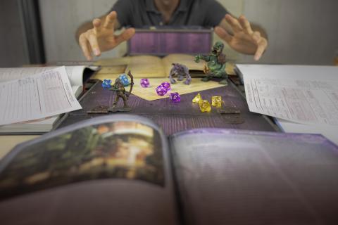 Photo of hands rolling colorful dice surrounded by roleplaying game books and miniatures