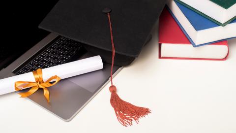 Diploma and graduation cap rest on a laptop computer with books next to it